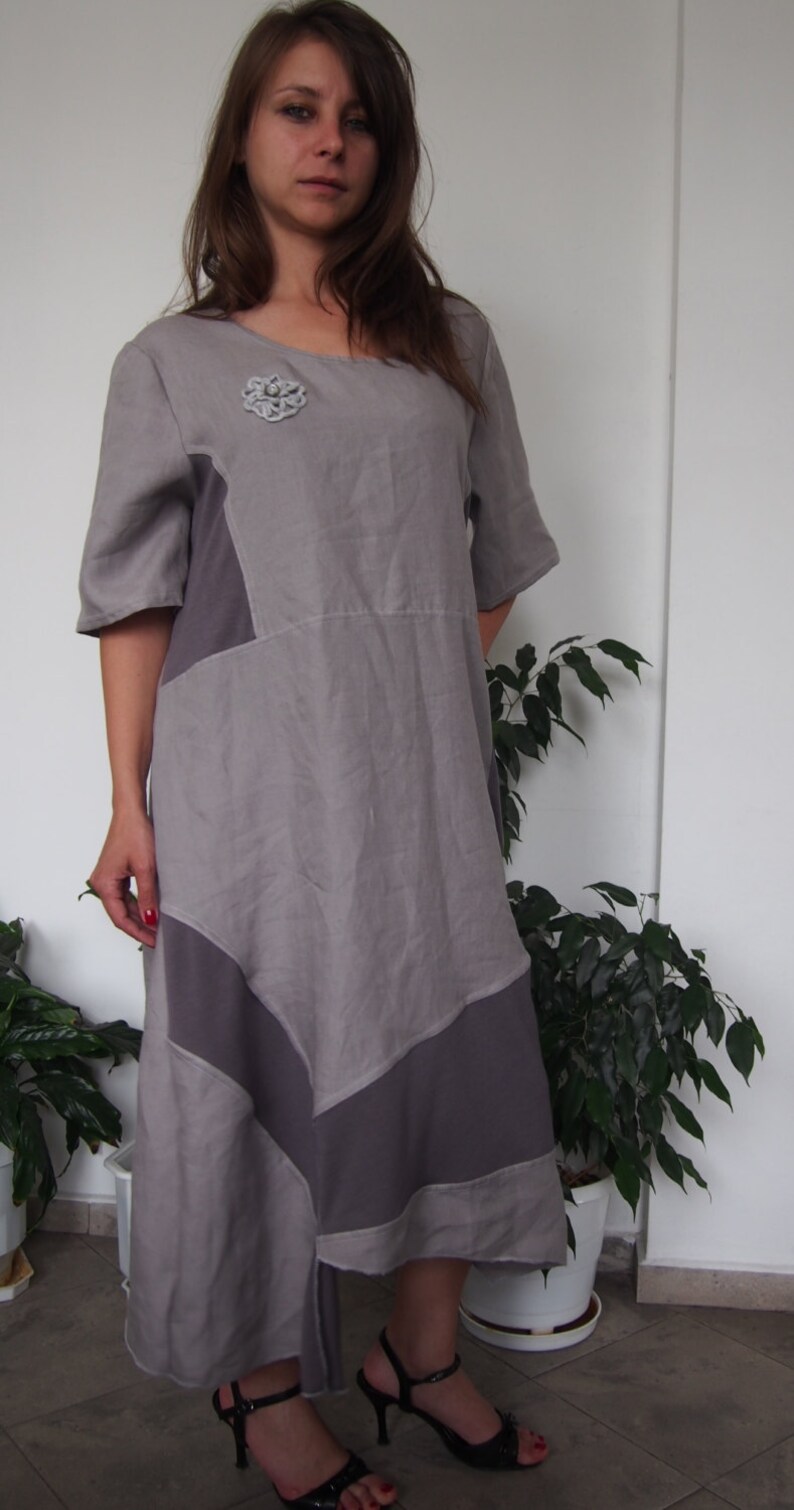May include: A gray linen dress with a geometric design and a silver flower brooch. The dress has a loose fit and a flowing skirt.