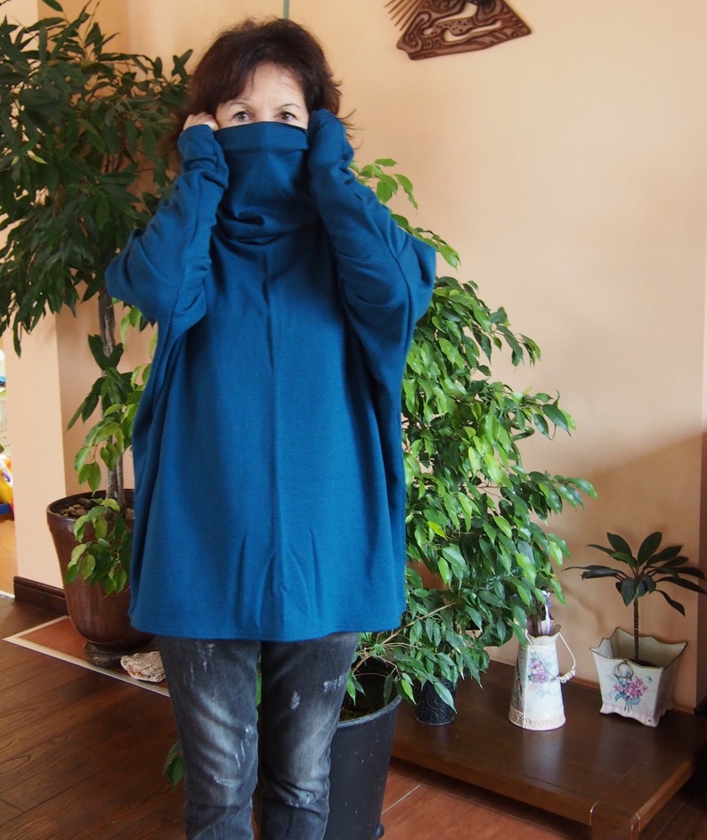 May include: A woman wearing a teal blue oversized turtleneck sweater with long sleeves and a high neckline. She is wearing blue jeans and is standing in front of a potted plant.