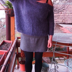 May include: A purple, fuzzy, knitted sweater with a turtleneck and a single, cut-out shoulder. The sweater is worn over a gray skirt and black tights.