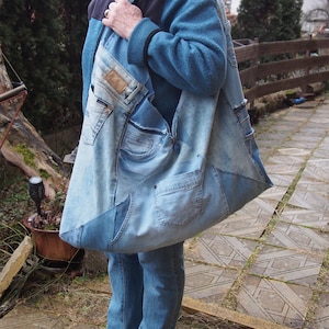 Puede incluir: Un bolso tote grande de mezclilla patchwork con múltiples bolsillos y una correa para el hombro. El bolso está hecho de jeans azules reciclados, mostrando varios tonos de mezclilla. El bolso es llevado por una persona que usa jeans azules y una chaqueta azul.