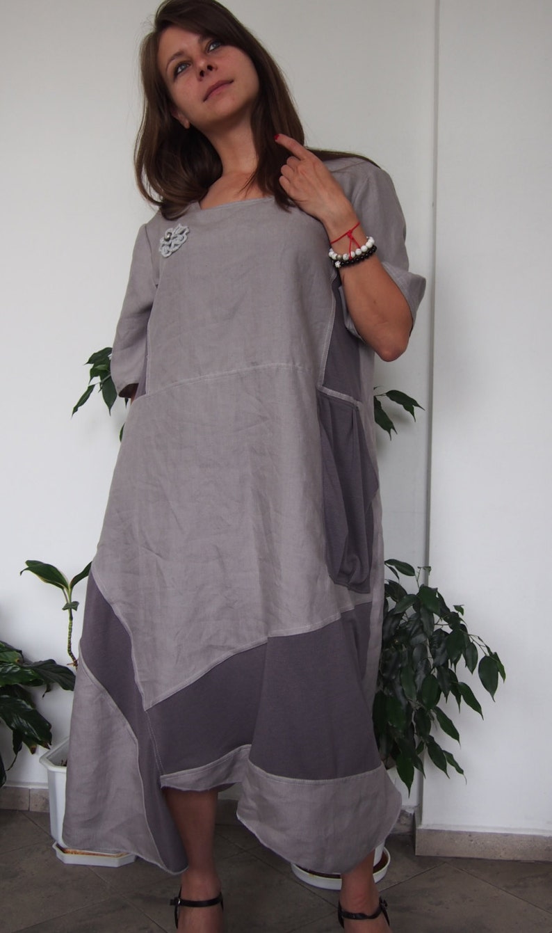 May include: A gray linen dress with a patchwork design. The dress has a scoop neck and short sleeves. It features a decorative flower brooch on the left side of the chest.