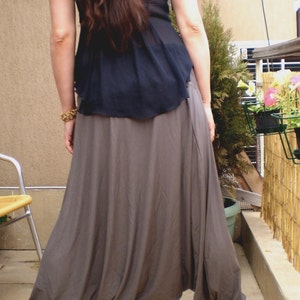 Puede incluir: Una mujer con un top negro y una falda larga, gris y fluida con un diseño fruncido en la parte inferior. La falda es suelta y ondeante, creando una silueta dramática. La mujer está de pie sobre un suelo de baldosas.