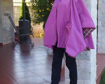 May include: A woman wearing a pink hooded poncho with a plaid lining. The poncho has a large pocket on the front. She is wearing black pants and black shoes.
