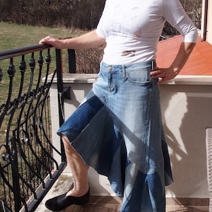 Op de afbeelding: Een vrouw draagt een wit T-shirt en een denim rok gemaakt van verschillende stukken denim. Ze staat op een balkon en houdt zich vast aan een zwart metalen hek.