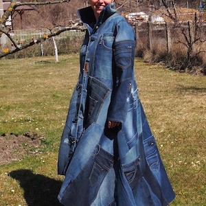 Op de afbeelding: Een lange patchwork denim jas in verschillende blauwtinten. De jas heeft meerdere zakken en een hoge kraag. Het kledingstuk lijkt handgemaakt en heeft een uniek ontwerp. De jas wordt buiten gedragen in een grasveld.