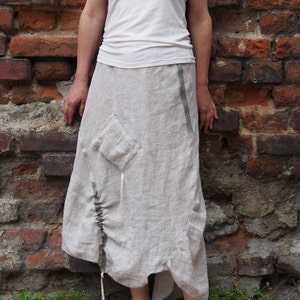 May include: A woman wearing a white tank top and a long, light beige linen skirt with a gathered detail and a side pocket. The skirt has a thin, olive green ribbon detail along the side seam. She is wearing silver sandals with red nail polish.