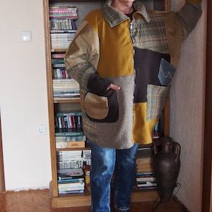 May include: A woman wearing a brown, yellow, and black patchwork jacket with blue jeans and brown boots. She is standing in front of a bookshelf filled with books.