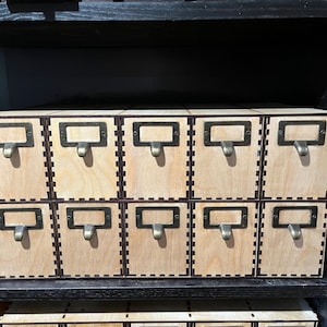 May include: A wooden box with twelve small drawers, each with a brass label holder and a metal pull. The drawers are arranged in two rows of six. The box is made of light wood with a dark brown stain.
