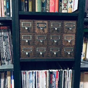 May include: A black wooden bookcase with multiple shelves filled with books and magazines. The middle shelf features a set of 12 small wooden drawers with brass pulls.