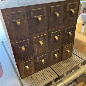 May include: A wooden box with twelve drawers, each with a brass pull. The box is made of dark wood and has a rustic look.