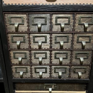 May include: A black wooden cabinet with 16 small drawers. Each drawer has a metal pull with a rectangular label.