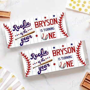 May include: Two baseball-themed candy bar wrappers with the text "Rookie of the Year" and "Bryson is turning one." The wrappers are white with red stitching and blue stars. Chocolate bars and decorative items surround the wrappers.