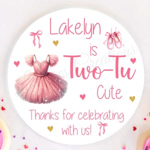 May include: A round, white sticker with pink text that reads "Lakelyn is Two-Tu Cute." The sticker features a pink tutu, ballet shoes, and hearts. Decorated cookies with pink frosting and sprinkles surround the sticker.