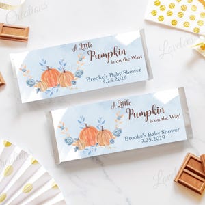 May include: Two baby shower candy bar wrappers with a watercolor design featuring pumpkins and floral accents. The wrappers read "A Little Pumpkin is on the Way!" and "Brooke's Baby Shower 9.25.2029". Decorative elements include chocolate bars and gold ribbon.