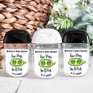 May include: Three clear hand sanitizer bottles with black and white lids. Each bottle has a label that reads "Brittney's Baby Shower Two Peas In A Pod 8-17-2022" with a graphic of two peas in a pod.
