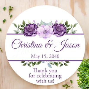 May include: Round white wedding sign with purple and light purple flowers, green leaves, and the text "Christina & Jason May 15, 2040 Thank you for celebrating with us!"