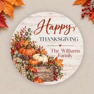 Puede incluir: Un letrero redondo de madera con fondo de veta de madera blanca que muestra el texto Happy THANKSGIVING y The Williams Family. El diseño incluye una caja llena de calabazas, piñas, hojas de otoño y un lazo de arpillera a cuadros.