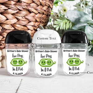 Puede incluir: Tres botellas de pl&aacute;stico transparente con tapas negras, blancas y negras. Cada botella tiene una etiqueta blanca con ilustraciones de guisantes verdes y el texto "Brittney's Baby Shower Two Peas In A Pod 6.15.2024". Una botella tiene el texto "Custom Text" sobre la etiqueta.