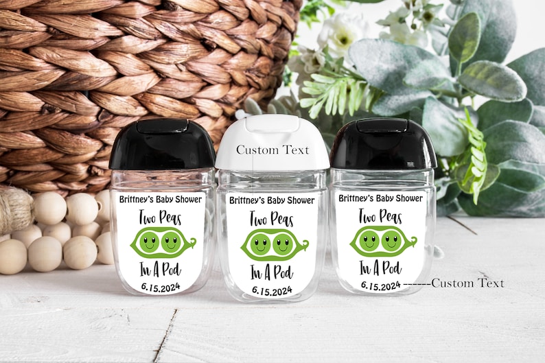 Puede incluir: Tres botellas de pl&aacute;stico transparente con tapas negras, blancas y negras. Cada botella tiene una etiqueta blanca con ilustraciones de guisantes verdes y el texto "Brittney's Baby Shower Two Peas In A Pod 6.15.2024". Una botella tiene el texto "Custom Text" sobre la etiqueta.