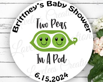 May include: A white round sticker with black text that reads "Brittney's Baby Shower" and "Two Peas In A Pod" with a green pea pod with two smiling peas inside. The date "6.15.2024" is also printed on the sticker.
