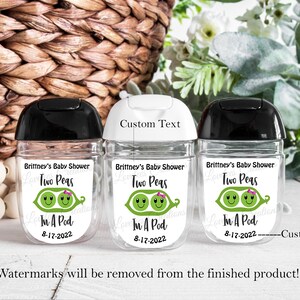 May include: Three clear, plastic hand sanitizer bottles with black caps. Each bottle has a label that reads "Brittney's Baby Shower Two Peas In A Pod 8-17-2022" with a graphic of two peas in a pod. The center bottle has the words "Custom Text" above it.