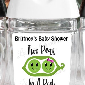May include: Clear, cylindrical hand sanitizer bottle with a white cap. The label reads "Brittney's Baby Shower" above a graphic of two smiling peas in a pod, with the text "Two Peas In A Pod" and the date "8-17-2022".