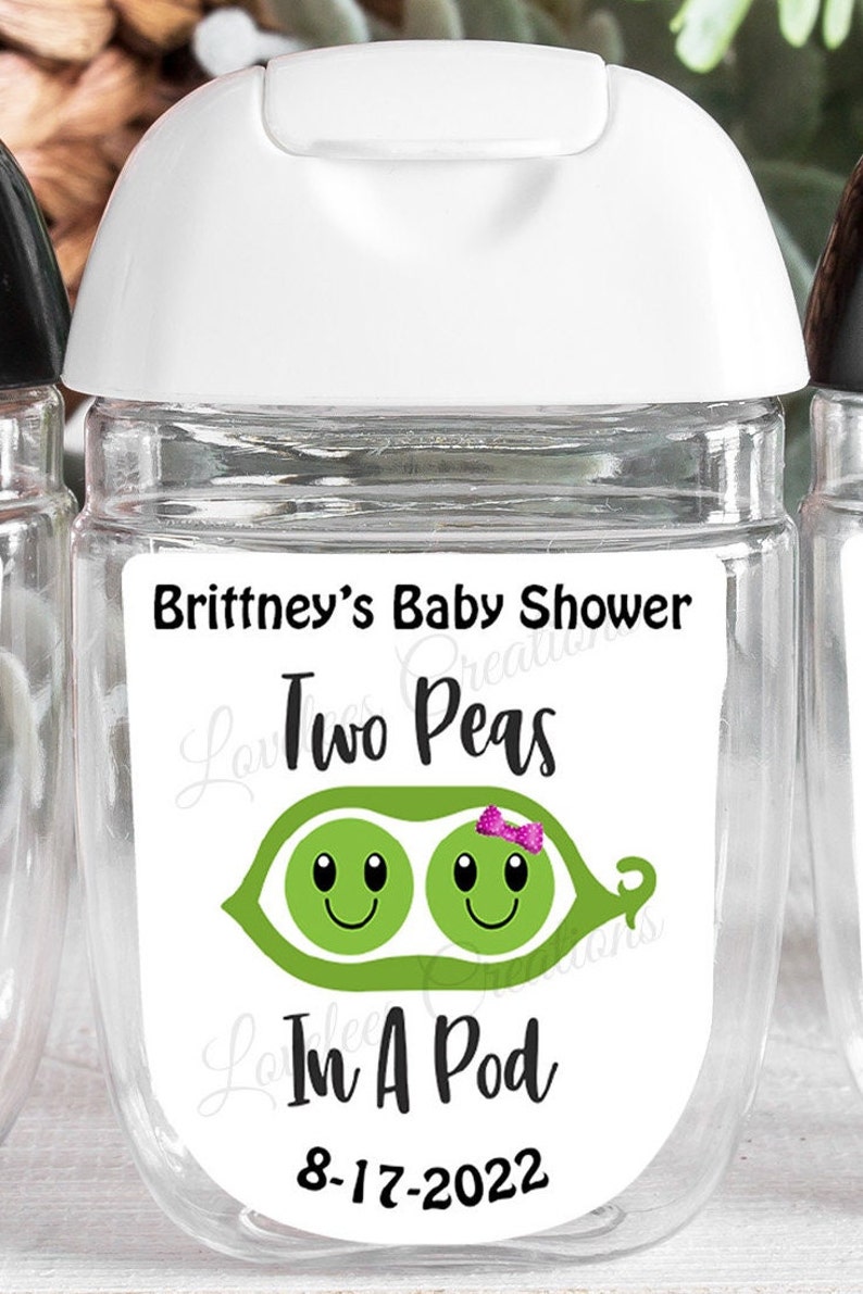 May include: Clear, cylindrical hand sanitizer bottle with a white cap. The label reads "Brittney's Baby Shower" above a graphic of two smiling peas in a pod, with the text "Two Peas In A Pod" and the date "8-17-2022".
