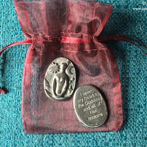 May include: Two silver metal tokens in a red drawstring bag. One token features a stylised image of a woman with a star and moon. The other token has the text "I open my heart to the Goddess and all of Her lessons".