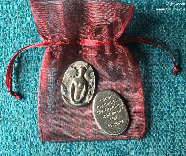 May include: Two silver metal tokens in a red drawstring bag. One token features a stylised image of a woman with a star and moon. The other token has the text "I open my heart to the Goddess and all of Her lessons".