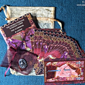 May include: A set of The Sacred Feminine Guidance Cards with a guidebook, a small pouch, and a card showing Guinevere the Queen. The cards have a red, gold, and purple colour scheme with intricate designs.