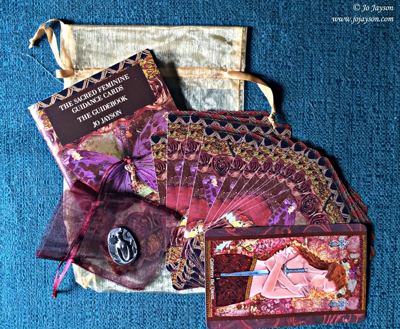 May include: A set of The Sacred Feminine Guidance Cards with a guidebook, a small pouch, and a card showing Guinevere the Queen. The cards have a red, gold, and purple colour scheme with intricate designs.