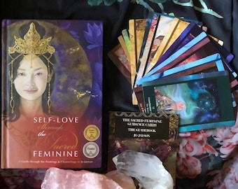 May include: A book titled Self-Love Through the Sacred Feminine by Jo Jayson, a deck of colorful oracle cards, a guidebook titled The Sacred Feminine Guidance Cards, and two raw crystals. The book cover features an illustration of a person wearing a gold crown.