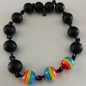 May include: A black beaded bracelet with alternating matte black and rainbow-striped beads. The bracelet is strung on a black chain and features a mix of solid and colorful spherical beads.
