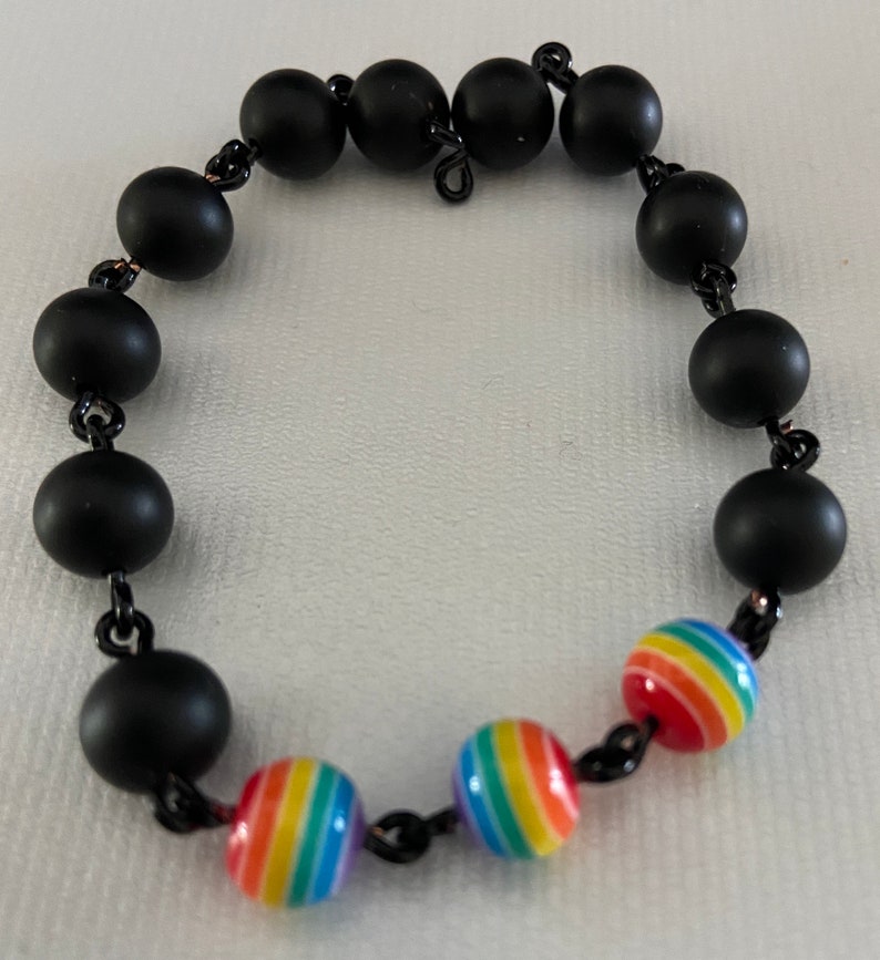 May include: A black beaded bracelet with alternating matte black and rainbow-striped beads. The bracelet is strung on a black chain and features a mix of solid and colorful spherical beads.