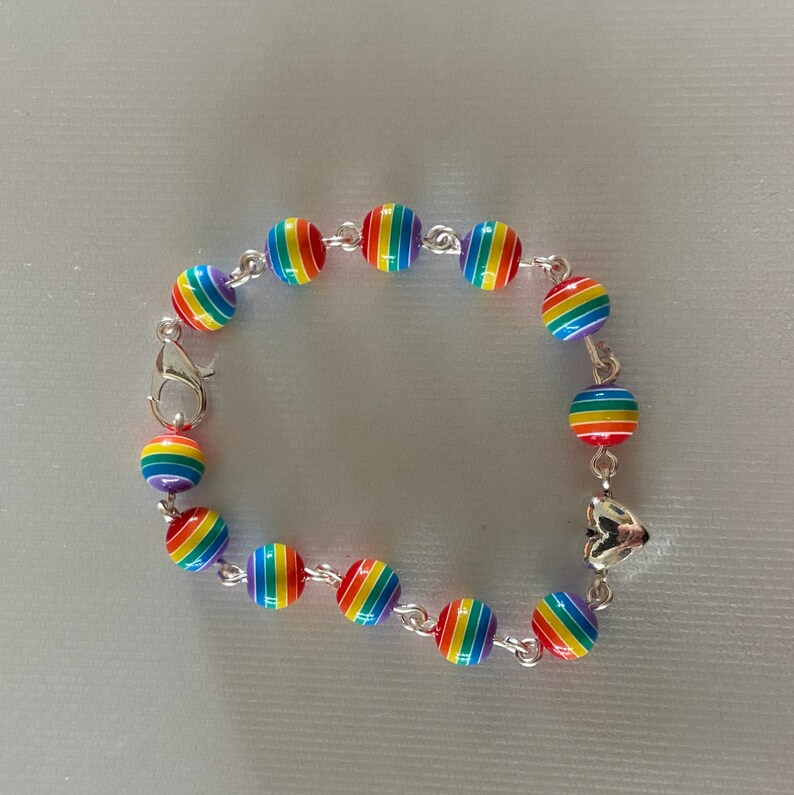 May include: A rainbow-striped bead bracelet with a silver clasp and connecting chain. The beads are round and feature a vibrant spectrum of colors. The bracelet is designed with a heart-shaped charm.