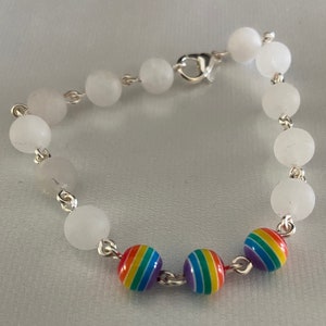 May include: A bracelet featuring white, round beads strung on a silver chain. The bracelet includes three rainbow-colored beads with stripes. The bracelet is secured with a silver clasp.