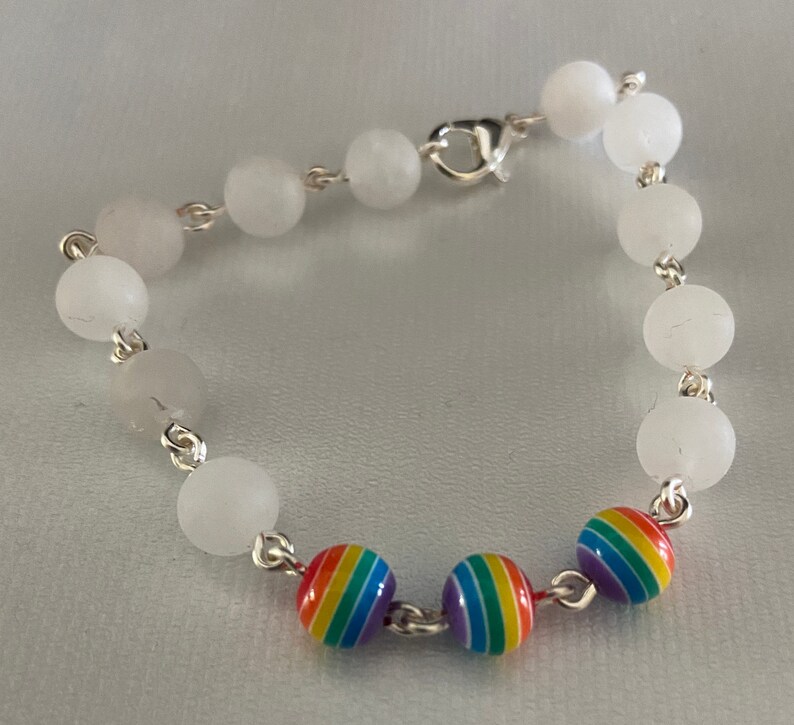 May include: A bracelet featuring white, round beads strung on a silver chain. The bracelet includes three rainbow-colored beads with stripes. The bracelet is secured with a silver clasp.