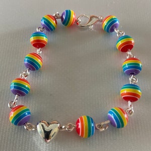 May include: A rainbow-colored beaded bracelet with a silver heart charm and clasp. The bracelet features round beads with horizontal stripes in red, orange, yellow, green, blue, and purple. The beads are strung together with a silver chain.
