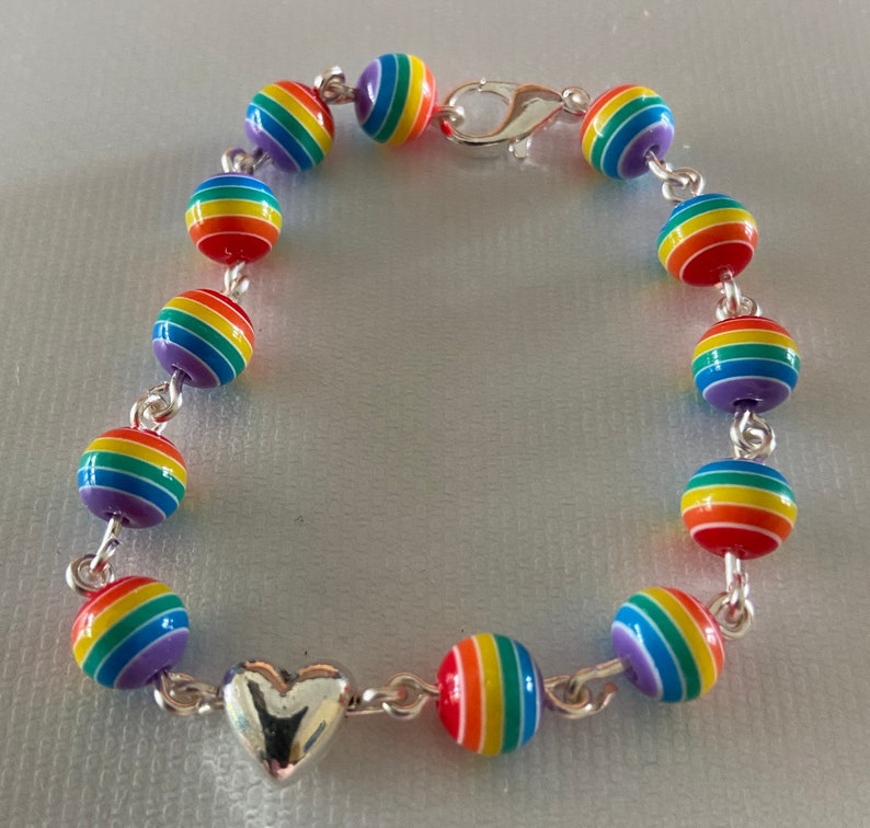 May include: A rainbow-colored beaded bracelet with a silver heart charm and clasp. The bracelet features round beads with horizontal stripes in red, orange, yellow, green, blue, and purple. The beads are strung together with a silver chain.
