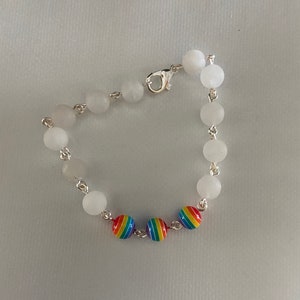 May include: A bracelet featuring white, round beads strung on a silver chain. The bracelet includes three rainbow-colored beads. The clasp is silver. The bracelet is displayed on a white surface.