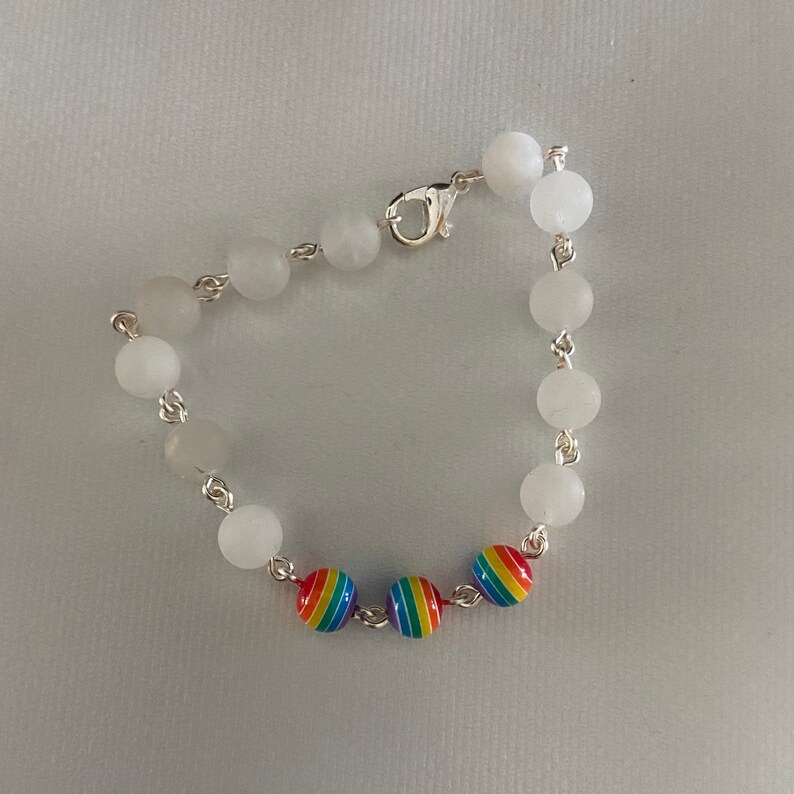 May include: A bracelet featuring white, round beads strung on a silver chain. The bracelet includes three rainbow-colored beads. The clasp is silver. The bracelet is displayed on a white surface.