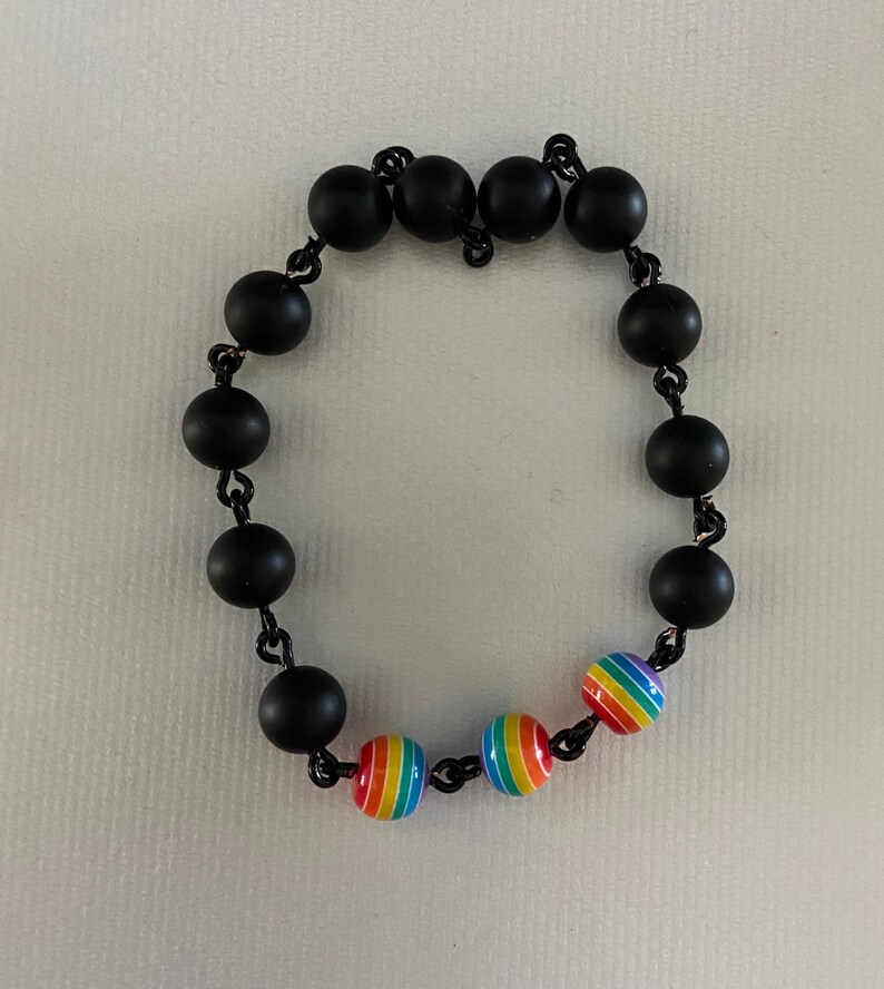 May include: A black beaded bracelet with rainbow-colored beads. The bracelet features matte black round beads strung together with a black chain. The rainbow beads have red, orange, yellow, green, blue, and purple stripes.
