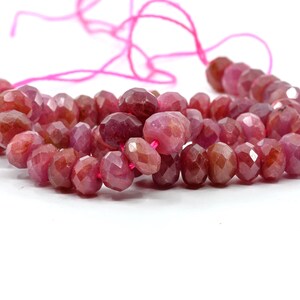 May include: A strand of faceted, round, translucent ruby-colored beads strung on a bright pink thread. The beads exhibit varying shades of deep red, magenta, and pink, with a polished, multi-faceted surface that reflects light across the strand.