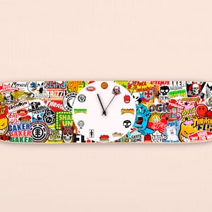 Puede incluir: Una esfera de reloj blanca con agujas negras está montada en una tabla de skate. La tabla está cubierta de pegatinas de marcas de skate de colores, incluyendo nombres como "Thrasher", "Santa Cruz", "Baker" y "Vans".