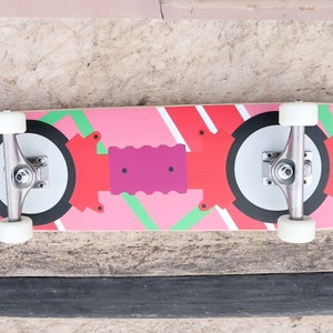 May include: A pink and red skateboard with black and white wheels. The deck has a geometric design with green accents.