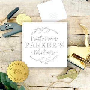 May include: A white square sign with the text "Fresh from Parker's Kitchen" in a decorative font, surrounded by leaf illustrations. The image also includes a gold-colored paper crimper and a lifetime guarantee seal.