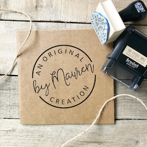 Puede incluir: Una tarjeta de papel kraft con un logotipo circular negro que dice "AN ORIGINAL by Mayren CREATION". Un sello de madera con un diseño floral azul y un sello autoentintable negro están colocados sobre una superficie de madera. También se ve hilo natural.