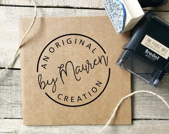 May include: A kraft paper card with a circular logo that reads "AN ORIGINAL by Mayren CREATION" in black script. A wooden stamp with a blue floral design and a black self-inking stamp are next to the card. The background is a rustic wooden surface.