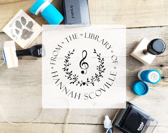 May include: A collection of stamps and ink pads on a wooden surface. The central stamp features a musical note and the text "FROM THE LIBRARY OF HANNAH SCOVILLE". Other stamps include a paw print and the name "Minnie".