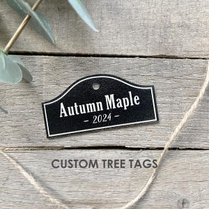 May include: A black, arched tree tag with the words "Autumn Maple - 2024" in white. The tag has a white border and a hole at the top. The words "CUSTOM TREE TAGS" are at the bottom.